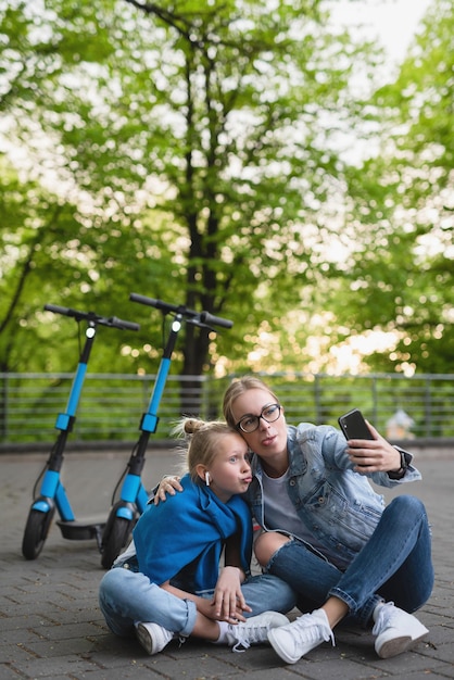 Felice madre e figlia che si fanno selfie mentre gli scooter elettrici viaggiano in un parco cittadino