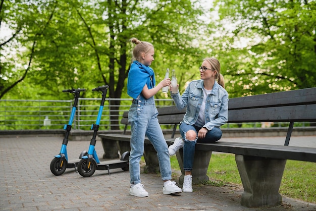 Felice madre e figlia con scooter elettrici stanno bevendo limonata nel parco cittadino