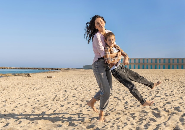 Felice madre e figlio che corrono e giocano sulla spiaggia a piedi nudi in una giornata di sole contro il faro