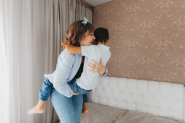 Felice madre e figlio piccolo stanno saltando sul letto, tenendosi per mano, ridendo la mamma sta giocando con il bambino in camera da letto.