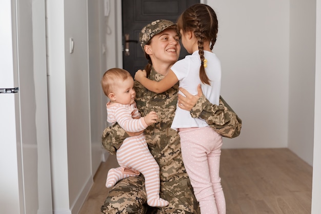 Felice madre militare soddisfatta che indossa l'uniforme e il cappello mimetici, tornando a casa dopo l'esercito e abbracciando i suoi figli, esprimendo emozioni positive, la femmina ha servito nell'esercito.