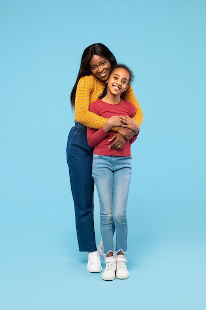 Felice madre nera che abbraccia la sua adorabile figlia in piedi in studio su sfondo blu a tutta lunghezza