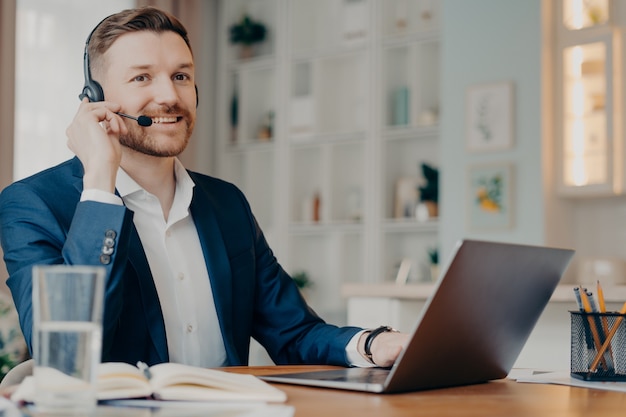 Felice manager maschio in tuta che indossa le cuffie e guarda mentre lavora con il laptop e parla online, uomo d'affari che ha riunioni online mentre lavora da remoto a casa. Concetto di lavoro a distanza