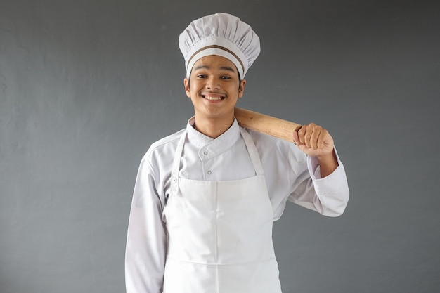 Felice maschio asiatico chef o panettiere in toque con mattarello su sfondo grigio