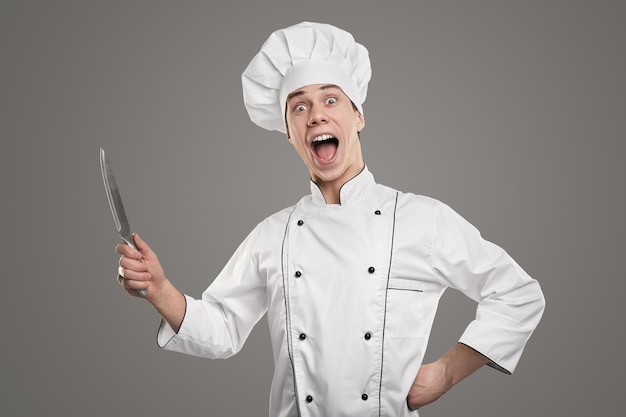 Felice maschio divertente in uniforme bianca da chef e con coltello che guarda la macchina fotografica pronta per preparare il cibo mentre guarda la macchina fotografica su sfondo grigio