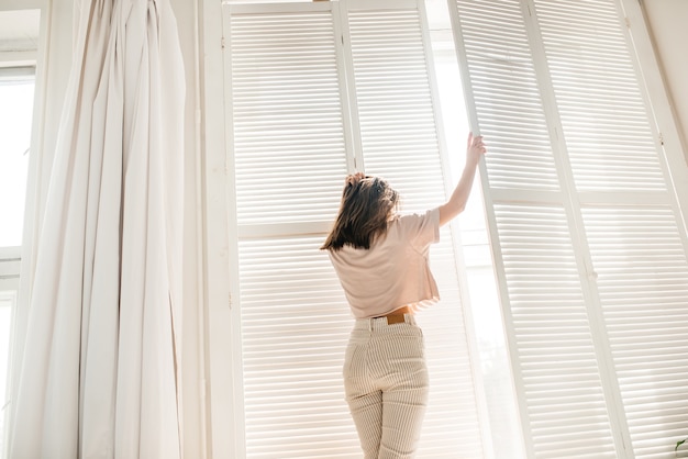 Felice mattina ragazza alla finestra dell'hotel, persiane bianche, luce solare