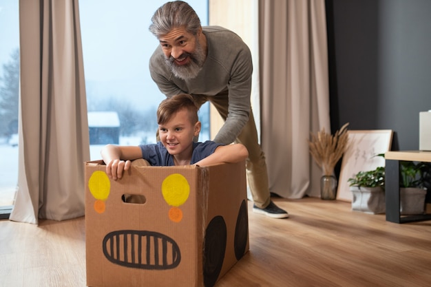 Felice maturo uomo barbuto in abbigliamento casual che spinge una scatola di cartone con suo nipote mentre entrambi si divertono durante il gioco in soggiorno