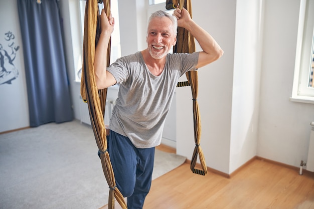 Felice maturo yogi caucasico dai capelli grigi che si aggrappa all'amaca yoga aerea durante l'allenamento