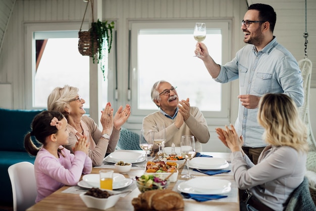 Felice metà uomo adulto tenendo un brindisi mentre pranza con la sua famiglia al tavolo da pranzo a casa