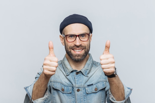 Felice modello maschile barbuto indossa occhiali, cappello elegante e giacca di jeans, mantiene i pollici sollevati