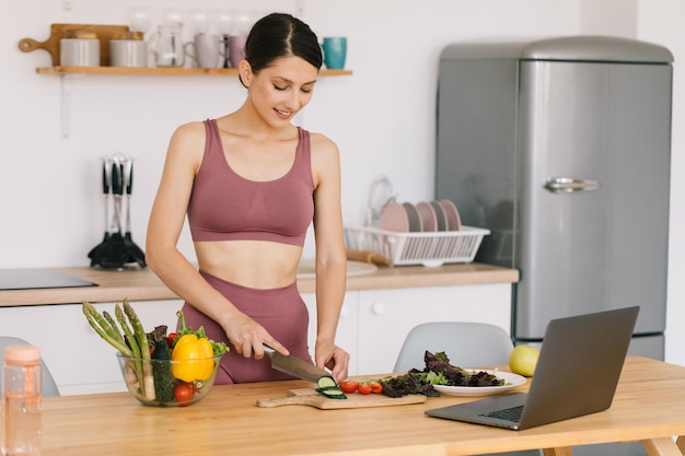 Felice nutrizionista blogger donna atletica prepara un'insalata con verdure fresche e conduce una videoconferenza su un'alimentazione sana sul laptop in cucina