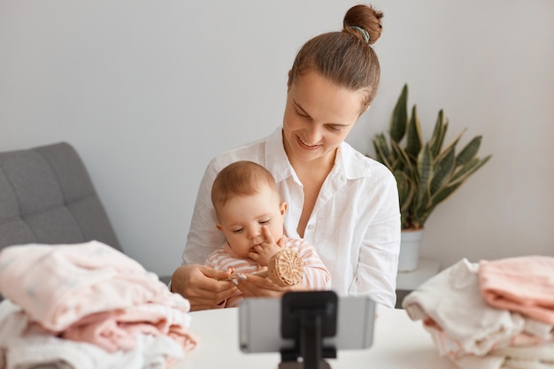 Felice ottimista madre caucasica seduta a tavola con un bambino piccolo e usa il telefono cellulare su un treppiede per trasmettere in livestream, creare contenuti per il suo blog.