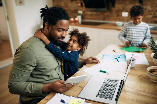 Felice padre afroamericano con due bambini che lavorano su laptop e touchpad a casa