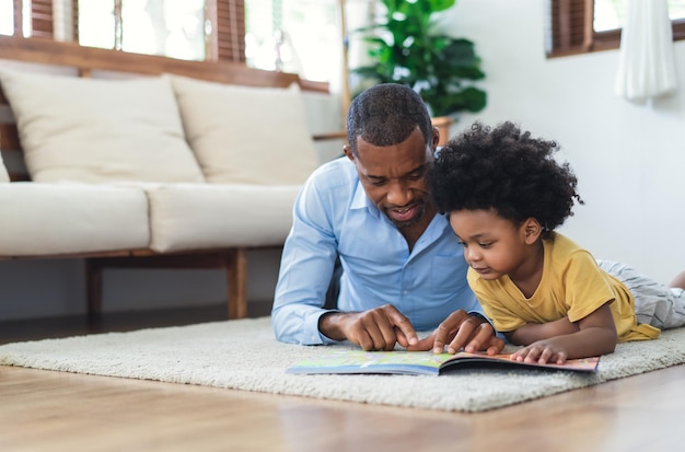 Felice padre e figlio afroamericano stanno leggendo un libro e sorridendo mentre giacevano sul pavimento trascorrendo del tempo insieme a casa Concetto di educazione e sviluppo dei bambini