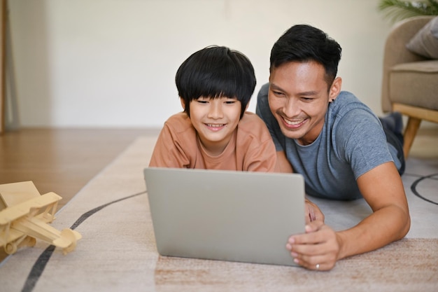 Felice papà asiatico è sdraiato sul pavimento a guardare un film o un cartone animato per bambini sul laptop con suo figlio