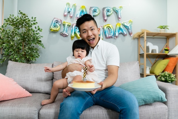 felice papà asiatico seduto sul divano sorride alla telecamera con una torta in mano e sua figlia in braccio mentre tiene la festa di compleanno nel soggiorno di casa.