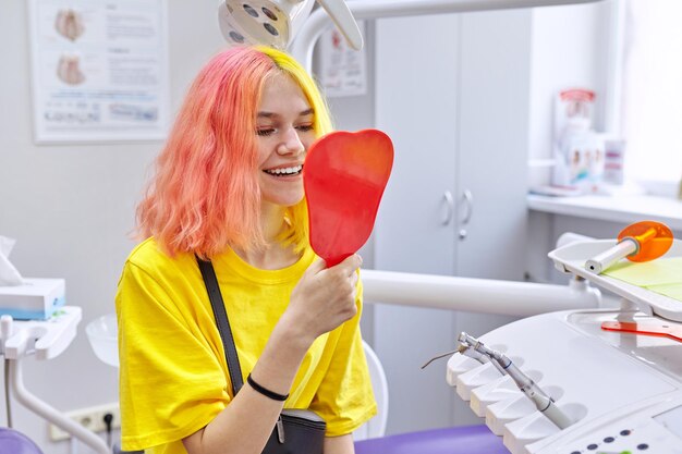Felice paziente bella ragazza adolescente guardando i suoi denti bianchi sani nello specchio sorridente mentre era seduto in poltrona odontoiatrica in clinica