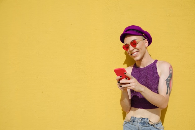 Felice persona non binaria che sorride mentre usa un telefono cellulare all'aperto contro un muro giallo. Tecnologia e concetto di stile di vita urbano.