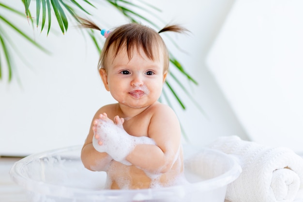 Felice piccola neonata si lava e gioca in una bacinella con schiuma e acqua in una stanza luminosa a casa