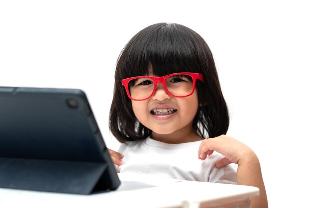 Felice piccola ragazza asiatica in età prescolare che indossa occhiali rossi e utilizza tablet pc su sfondo bianco e sorride, ragazza asiatica che impara con una videochiamata con tablet, concetto educativo per i bambini delle scuole