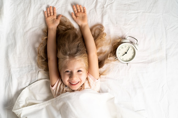 Felice piccola ragazza bionda in un letto bianco con una sveglia, buongiorno.