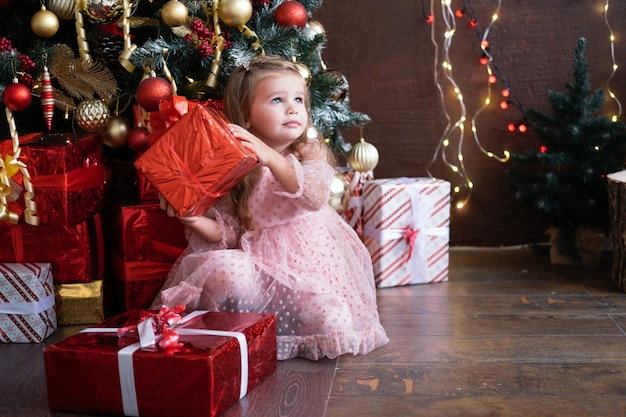 Felice piccola ragazza bionda sorridente in un bel vestito con scatola regalo vicino all'albero di natale