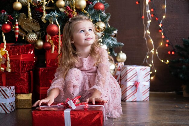 Felice piccola ragazza bionda sorridente in un bel vestito con scatola regalo vicino all'albero di natale