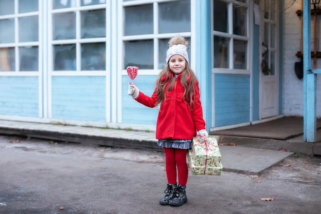Felice piccola ragazza con cappello bianco e cappotto rosso in piedi per strada con dolci caramelle lecca-lecca natalizie