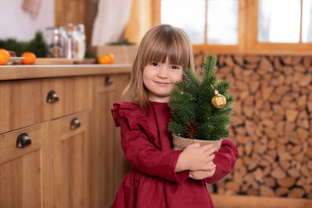 Felice piccola ragazza in abito rosso che mostra un piccolo abete di Natale decorato e sorridente
