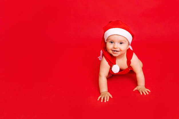 Felice piccolo bambino di sei mesi con cappello da Babbo Natale che striscia su un fotofono rosso isolato, spazio per il testo, il nuovo anno e il concetto di vacanza