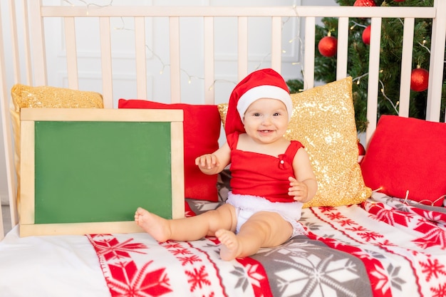 Felice piccolo bambino sei mesi in un cappello da Babbo Natale seduto in una culla a casa dall'albero di Natale con una scheda di testo, il nuovo anno e il concetto di vacanza