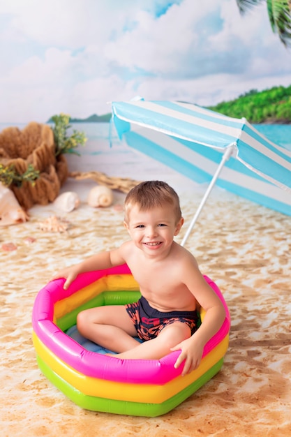 Felice piccolo neonato bagna in una luminosa piscina gonfiabile su una spiaggia di sabbia con palme in riva al mare