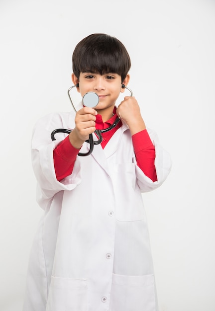 Felice piccolo ragazzo asiatico indiano in uniforme medica come medico, tenendo in mano uno stetoscopio e guardando la telecamera isolata su sfondo bianco white