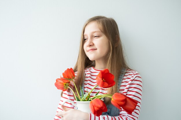 Felice ragazza adolescente con capelli lunghi che tiene vaso con tulipani rossi in piedi contro il muro bianco