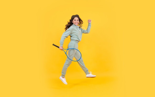 Felice ragazza adolescente energica salta in abbigliamento sportivo con racchetta da badminton che corre verso il successo su sfondo giallo sbrigati