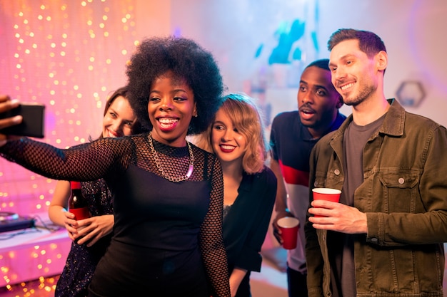 Felice ragazza africana con il sorriso a trentadue denti facendo selfie con amici interculturali mentre vi godete la festa a casa e bevete insieme