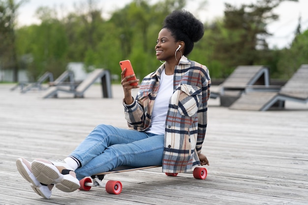 Felice ragazza africana tiene lo smartphone seduto nel parco urbano su longboard rilassato ascolta musica e messaggi