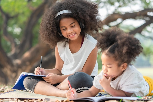 Felice ragazza afroamericana guarda sua sorella minore sdraiata disegno nel libro da colorare nel parco