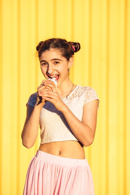 Felice ragazza alla moda in gonna rosa macchiata il naso con gelato e sorriso sul muro giallo