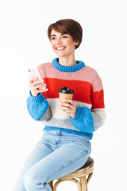 Felice ragazza allegra che indossa un maglione seduto su una sedia isolata su bianco, utilizzando il telefono cellulare, tenendo tazza da asporto