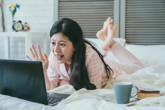 felice ragazza asiatica che indossa il pigiama sdraiato prono sta salutando la videocamera del computer mentre fa una chat video con un amico e fa colazione sul letto nella camera da letto a casa