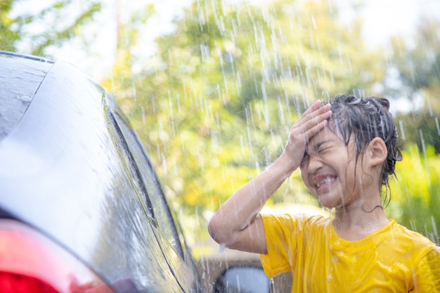 Felice ragazza asiatica che lava l'auto su spruzzi d'acqua e luce solare a casa