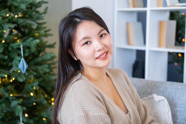 felice ragazza asiatica in cardigan beige con confezione regalo seduta sul divano a casa vicino all'albero di natale