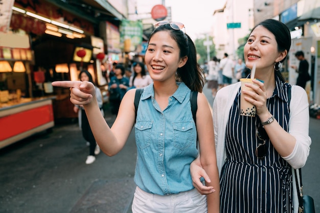 felice ragazza asiatica sta indicando a distanza e suggerendo alla sua amica con un tè al latte bolle di fare il check-out mentre fa shopping insieme in una strada turistica in estate