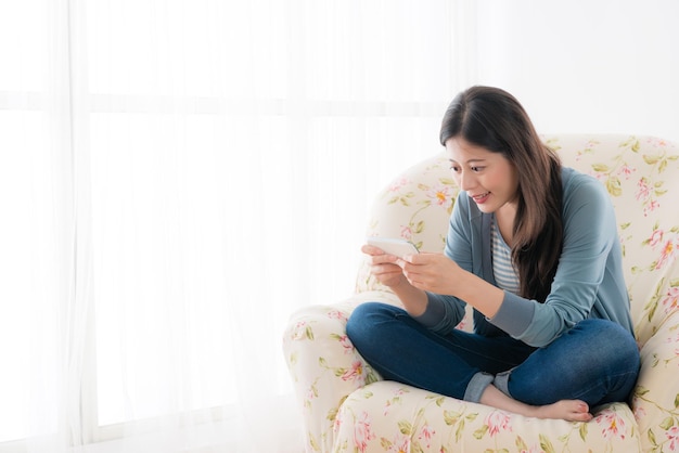 felice ragazza attraente seduta su un comodo divano davanti alla finestra del soggiorno e utilizzando il cellulare per giocare online.