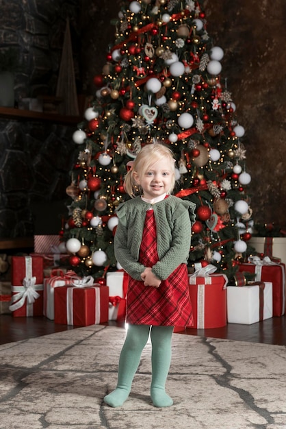 Felice ragazza bionda in abito rosso vicino albero di Natale Ritratto di bambino carino su sfondo albero di NataleTelaio verticale