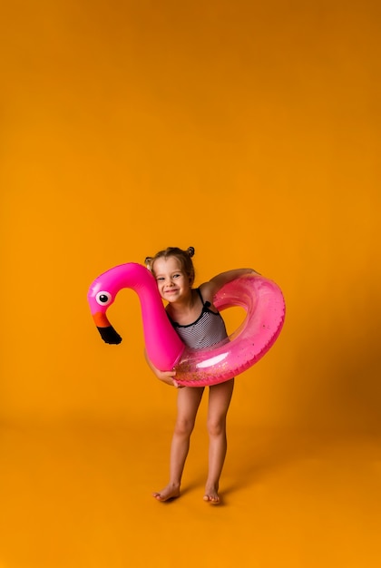 Felice ragazza bionda in un costume da bagno a righe tiene un cerchio gonfiabile rosa con un fenicottero su una superficie gialla con un posto per il testo