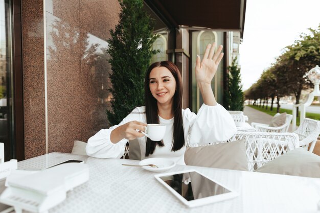 Felice ragazza bruna si siede in un caffè e agita la mano a un amico per strada.