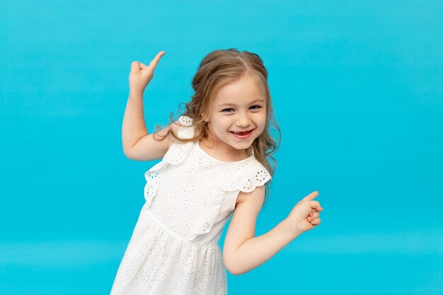 Felice ragazza carina in un abito bianco di cotone su sfondo blu in studio ridendo mostrando le sue dita nel ver, sorridendo e scherzando, un posto per il testo