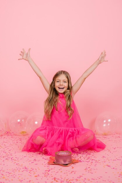 Felice ragazza caucasica sorridente di otto anni ha espresso un desiderio vicino a una torta bento per il suo compleanno ha alzato con gioia le mani ai lati seduta vicino a palloncini su uno sfondo rosa con coriandoli